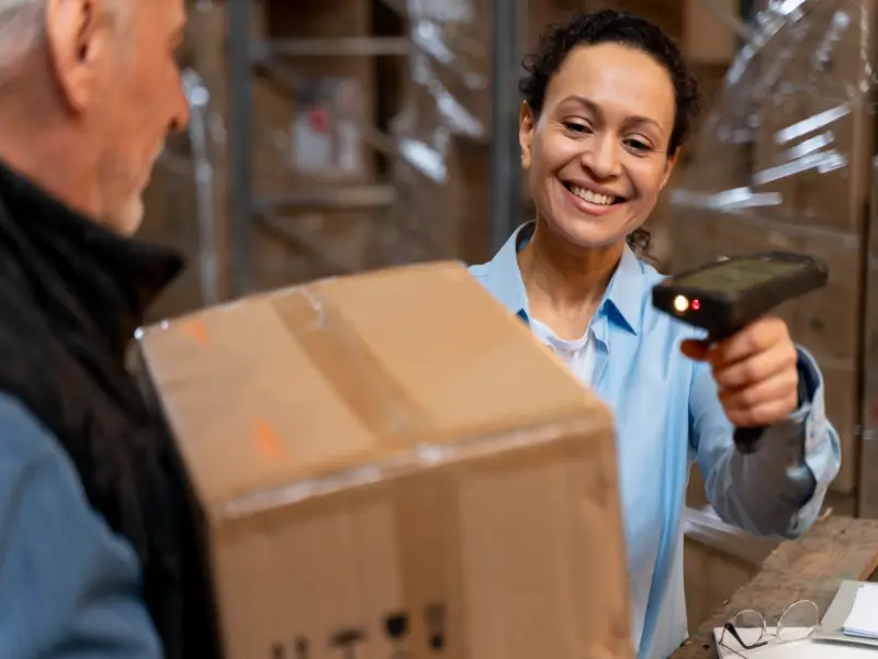 A warehouse employee uses a handheld barcode scanner to process a shipping box held by a worker, showcasing the inventory management and fulfillment side of a dropshipping business